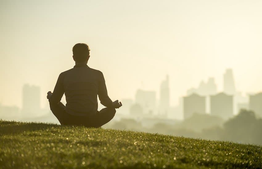 Silhouette of a man sitting in the lotus position meditating on the grassy top of Primrose Hill in front of a misty golden sunrise view of the London city skyline